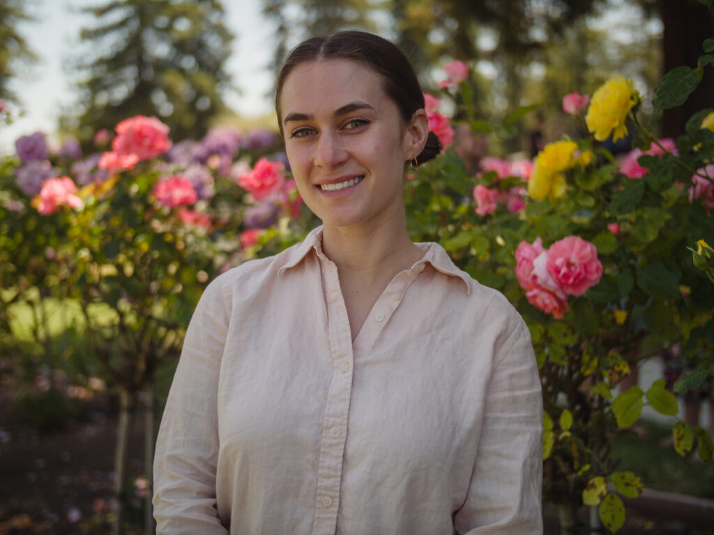 Haley Witt, founder of Blue Heron Therapy, standing in front of bushes that are flowering with yellow and pink flowers.